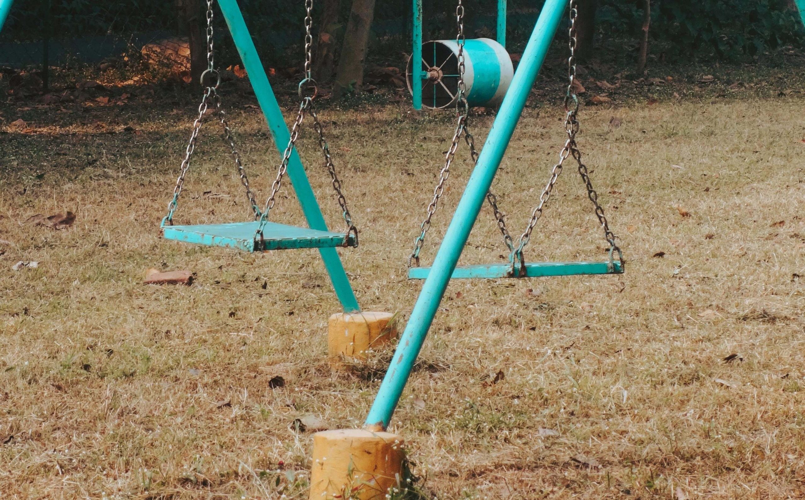 Two teal colored swings that appears to be rusting in the middle of an unkept park.