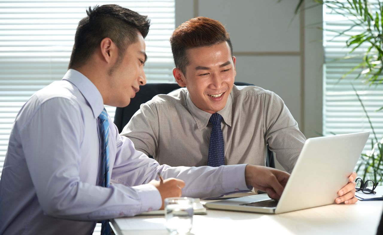 Two Asian men meet in an office as they look at a laptop together.