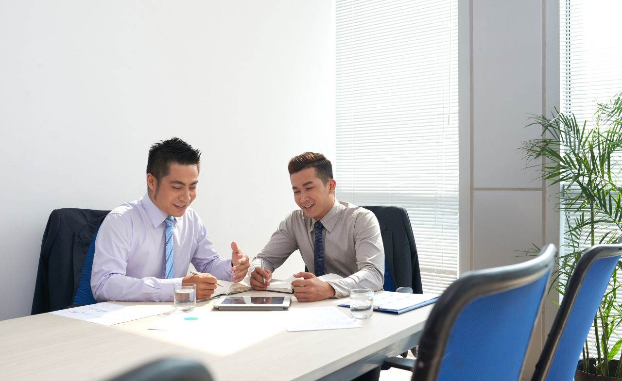 Two Asian men sit at a table in a meeting room, having a discussion.